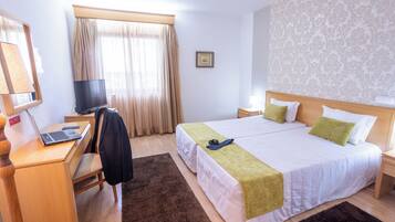 A hotel room with a bed, a desk with a laptop, a chair, a television, and a window with curtains.