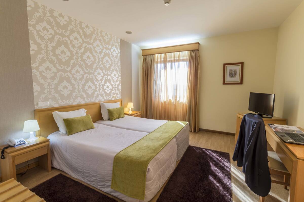Ein Hotelzimmer mit zwei Betten, einem Schreibtisch mit Computer, einem Fernseher und einem Fenster mit Vorhängen.