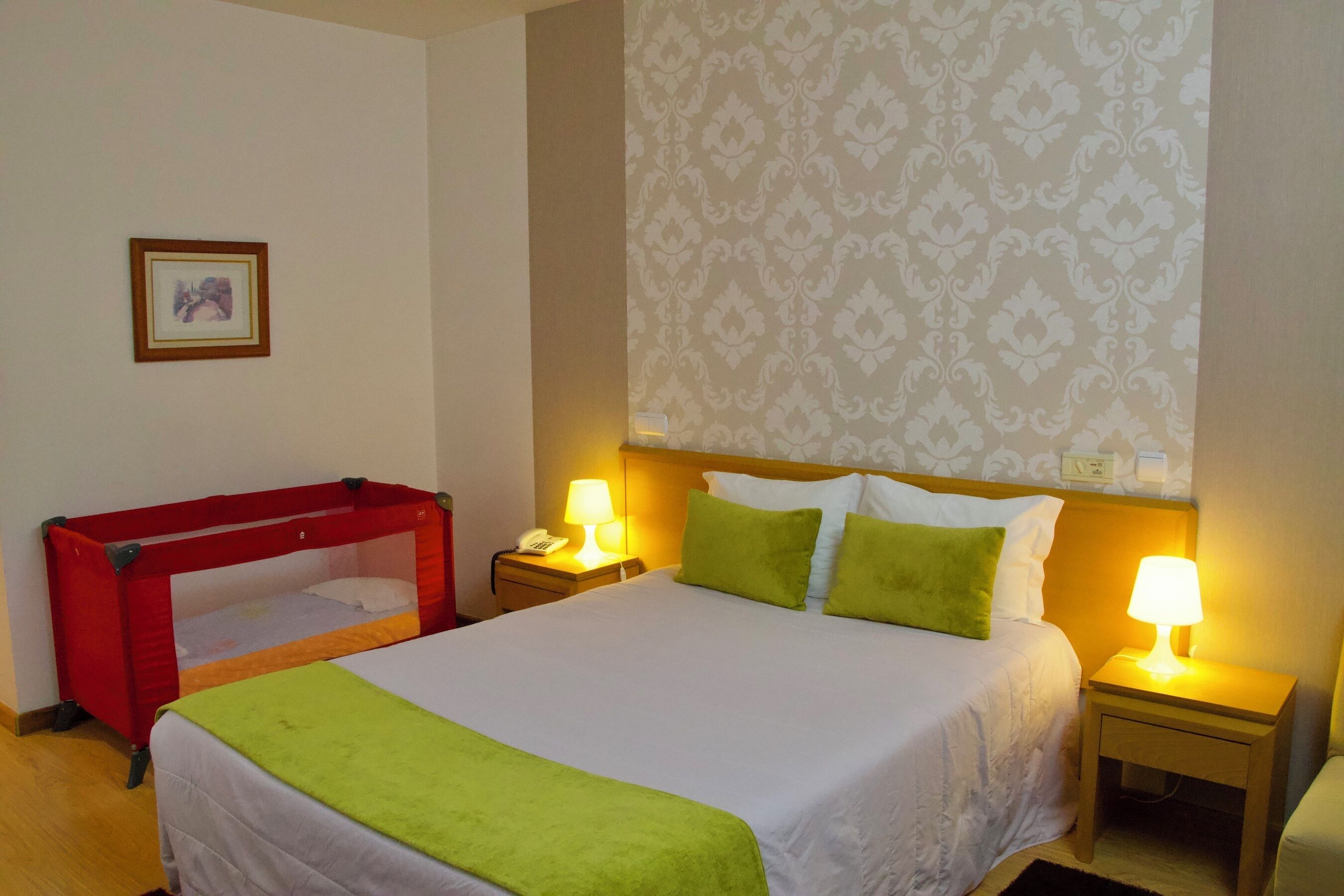Een hotelkamer met een eenpersoonsbed, twee bedlampen, een nachtkastje, een rode kinderbedje en behang met een patroon.