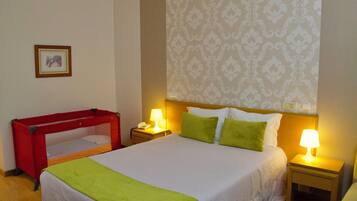 A hotel room with a single bed, two bedside lamps, a nightstand, a red crib, and a patterned wallpaper.