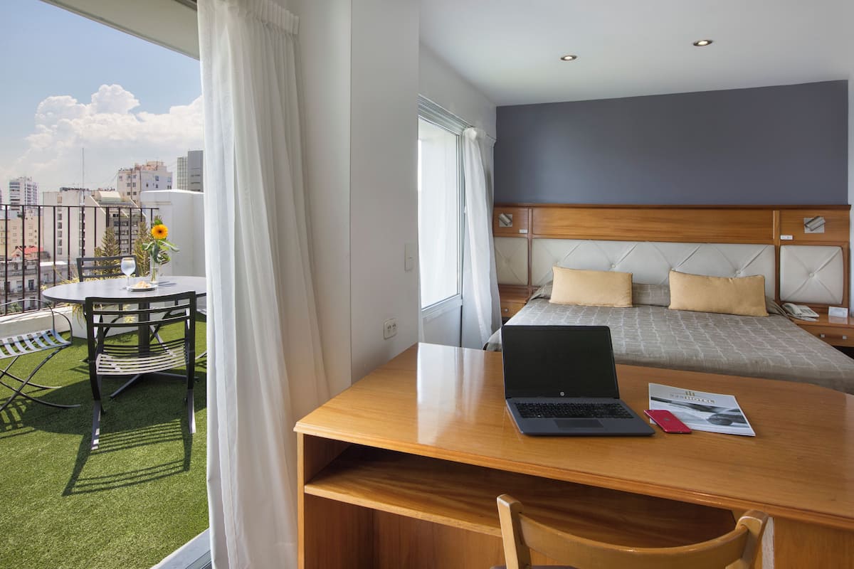A hotel room with a balcony, a bed with a wooden headboard, a desk with a laptop, and a view of the city.