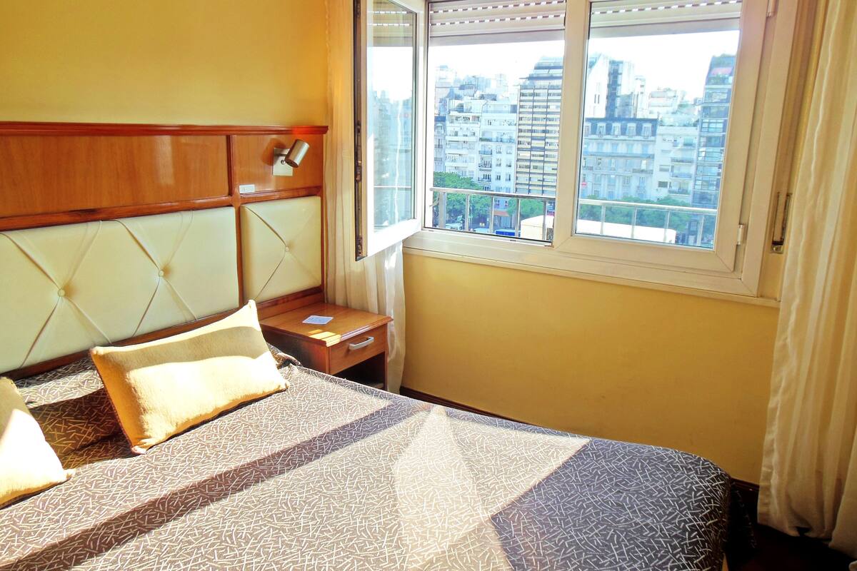 A hotel room with a bed, bedside table, lamp, and a window with a city view.