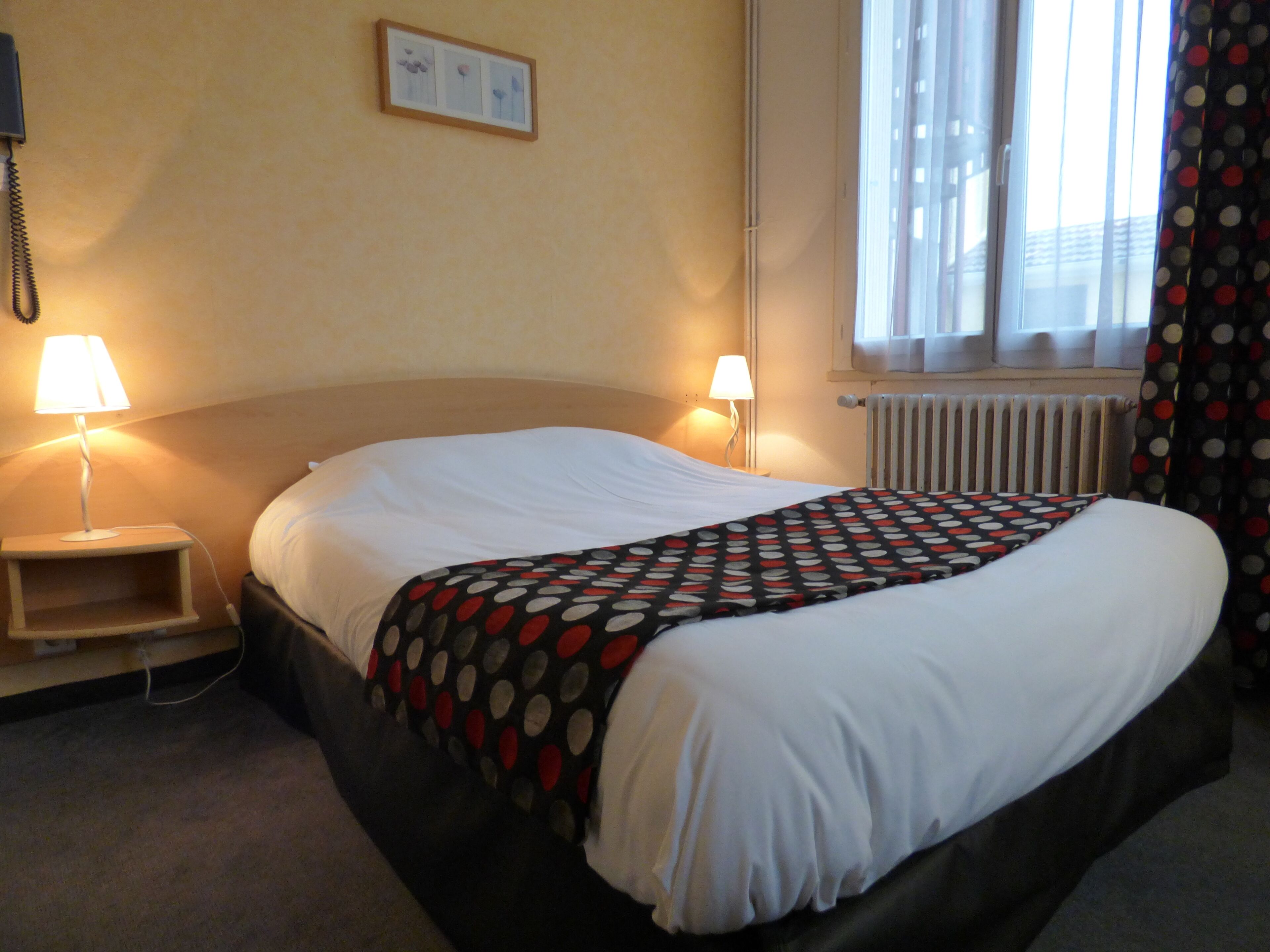 Une chambre d’hôtel avec un lit, deux lampes de chevet, un tableau encadré au mur, une fenêtre avec des rideaux et un radiateur.
