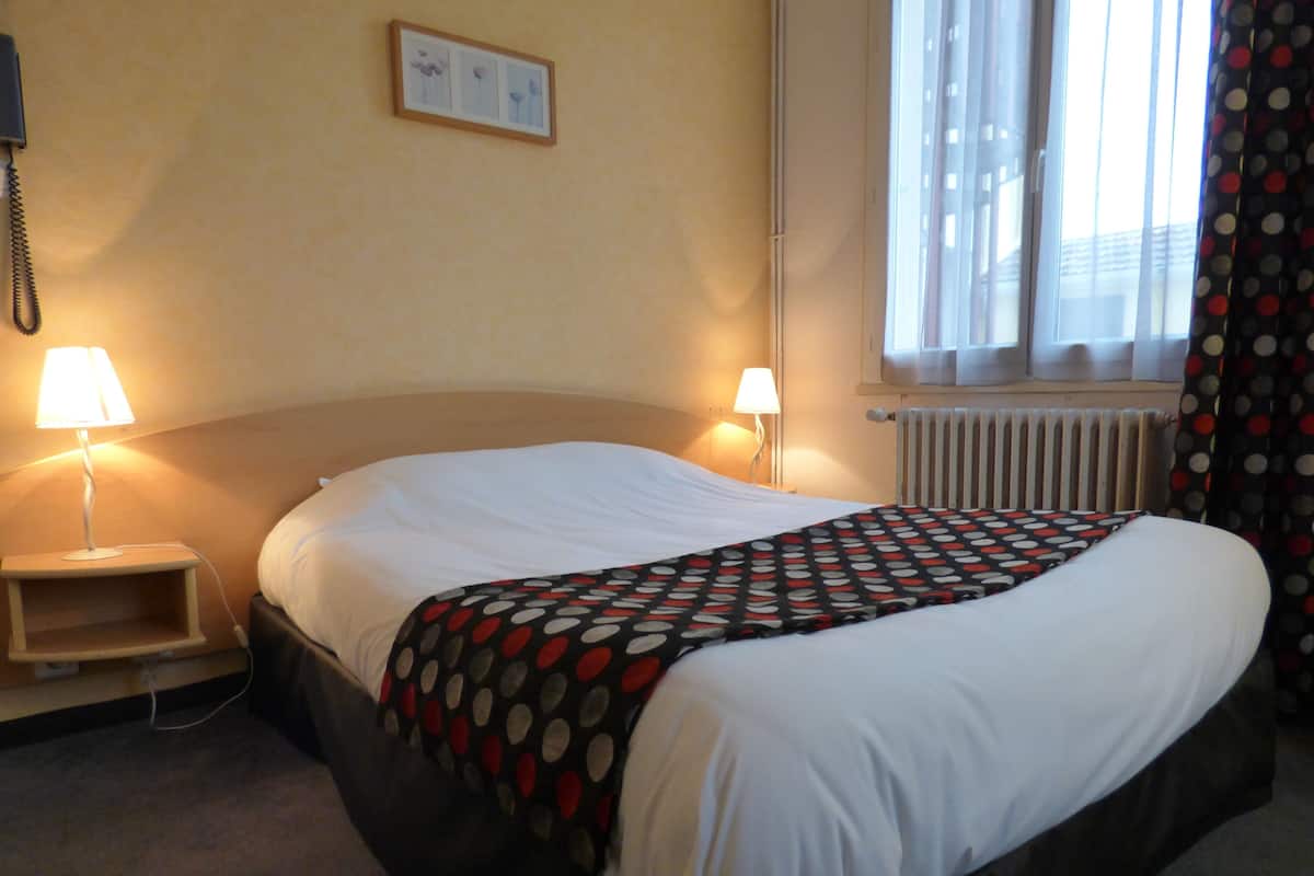 Une chambre d’hôtel avec un lit, deux lampes de chevet, un tableau encadré au mur, une fenêtre avec des rideaux et un radiateur.