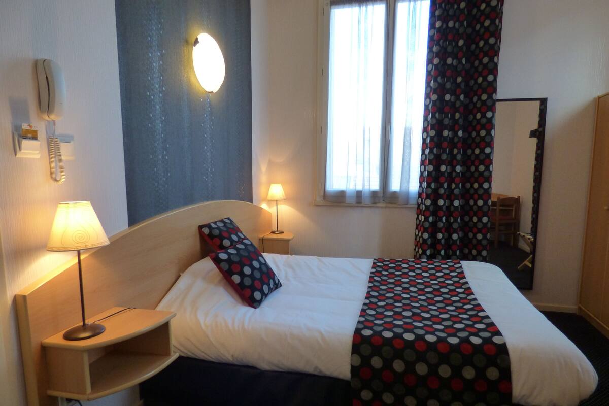 Une chambre d’hôtel avec un lit, deux lampes de chevet, un téléphone fixé au mur, une fenêtre avec des rideaux et un miroir.