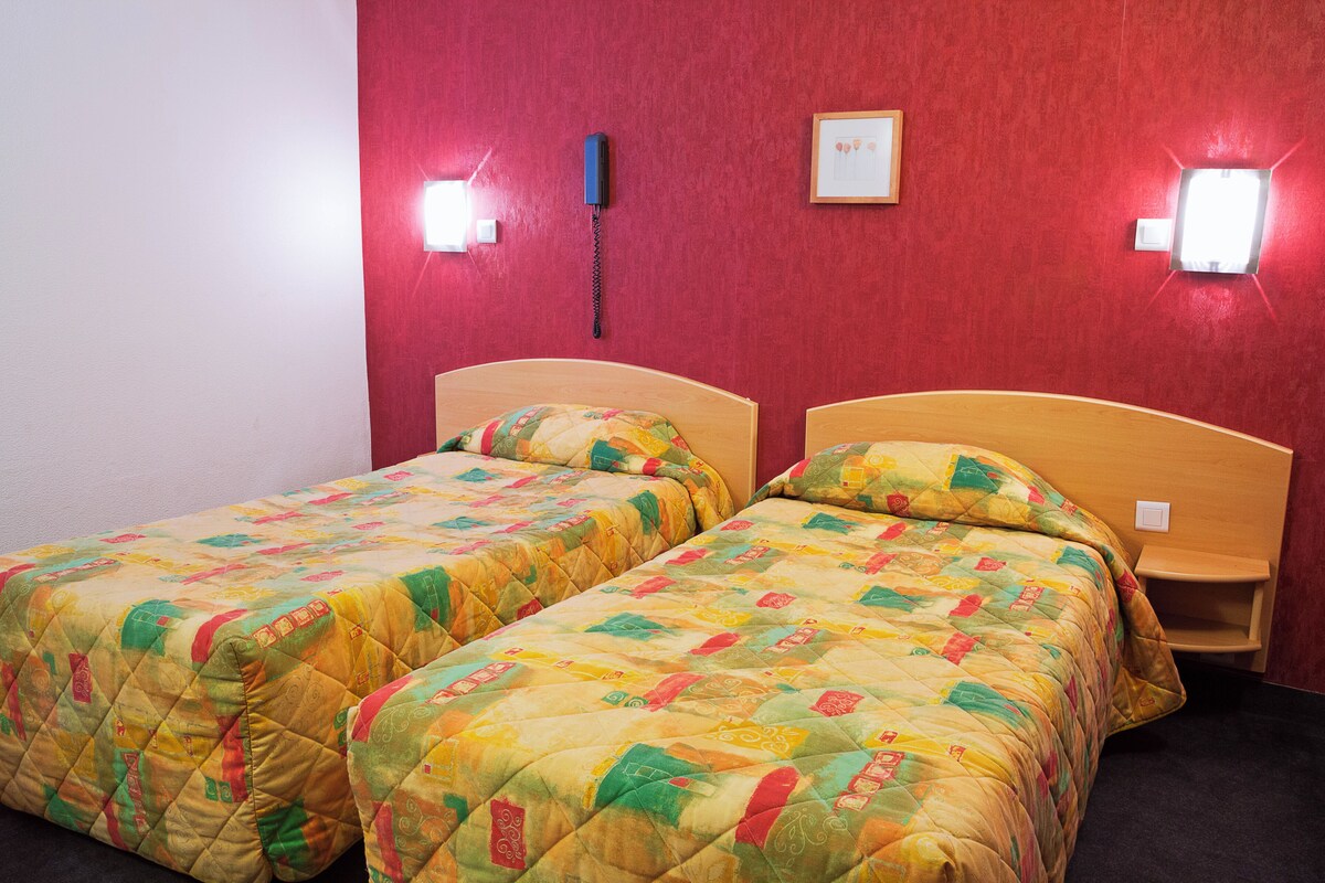 A room with two beds, each with a quilted bedspread and wooden headboards. There are two wall-mounted light fixtures and a telephone on the wall.