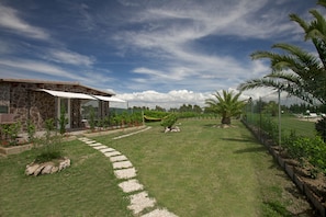 A stone building with a covered patio, a grassy lawn with stepping stones, and a fenced area with plants and a hammock. - Fattoria Maremmana (Grosseto)