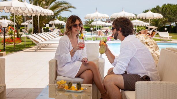 Una pareja sentada a una mesa con bebidas, con vista a un área de piscina con sillones y sombrillas.