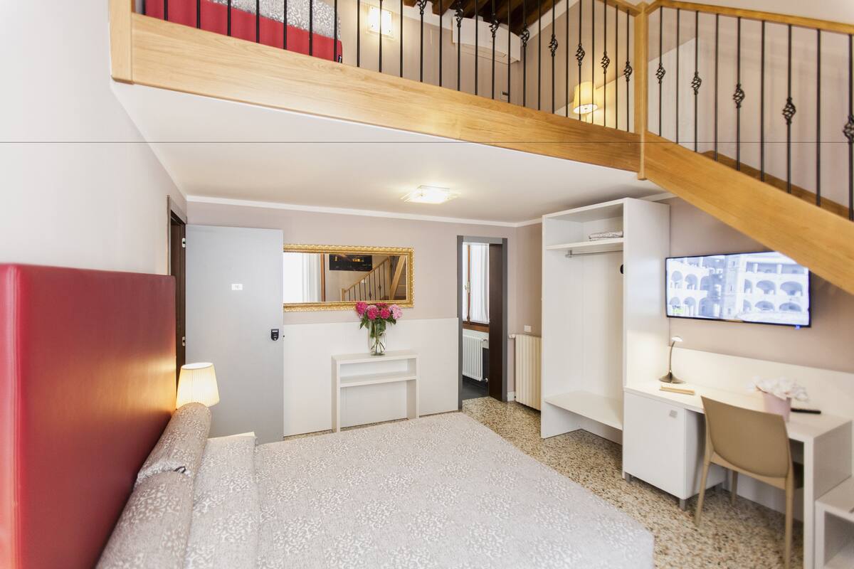 Habitación de hotel con cama, escritorio, silla, televisor, espejo y una escalera.