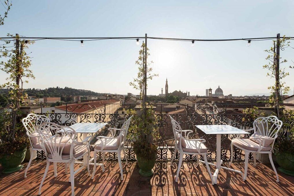 A terrace with white metal chairs and tables, overlooking a cityscape with historic buildings and a clock tower.