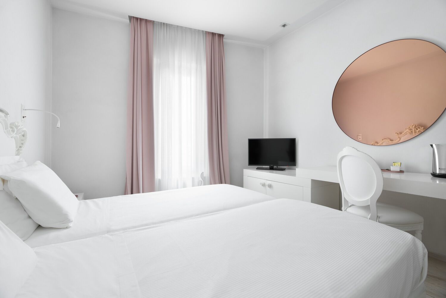 Habitación de hotel con una cama grande, un escritorio con televisor, una silla y un espejo redondo en la pared.