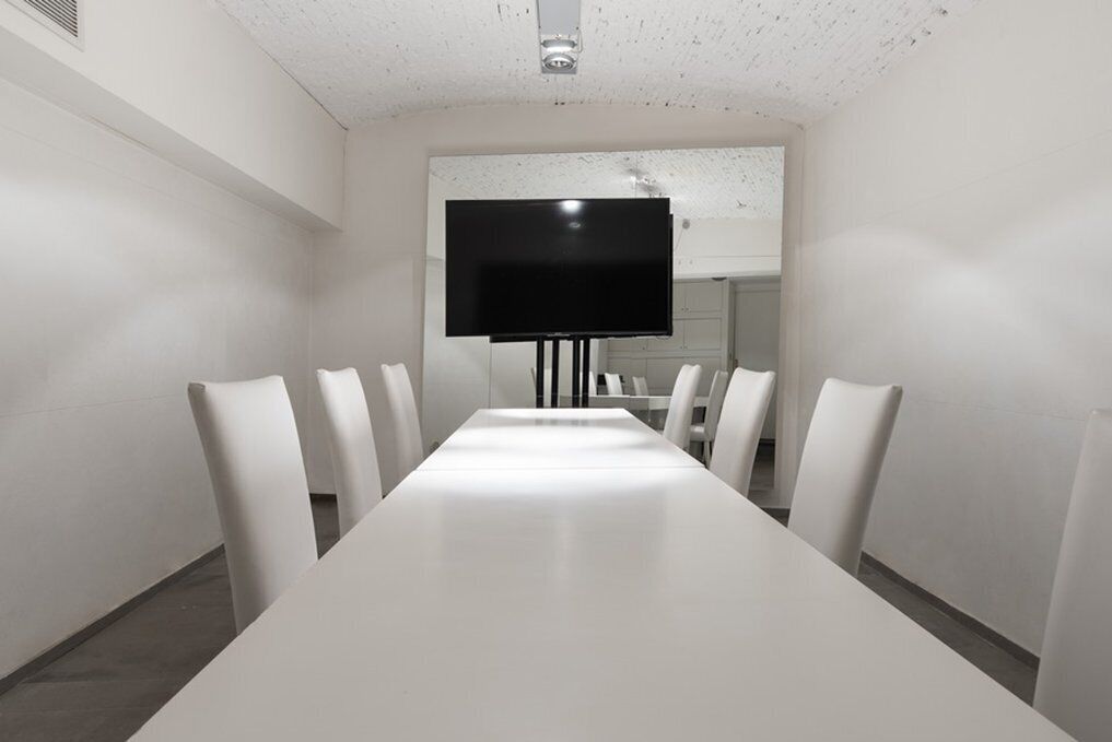 Un salón de conferencias con una larga mesa blanca, seis sillas blancas y un televisor de pantalla plana montado en la pared.
