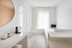 Camera d'albergo minimalista con un letto, una scrivania con bollitore, un tavolino e una TV.