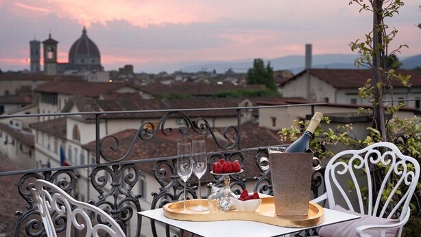 A balcony with a table set for two, featuring a bottle, glasses, and a tray with food and flowers.