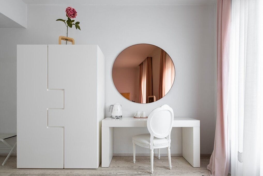 Una habitación minimalista con un escritorio blanco, una silla, un espejo redondo y un armario.