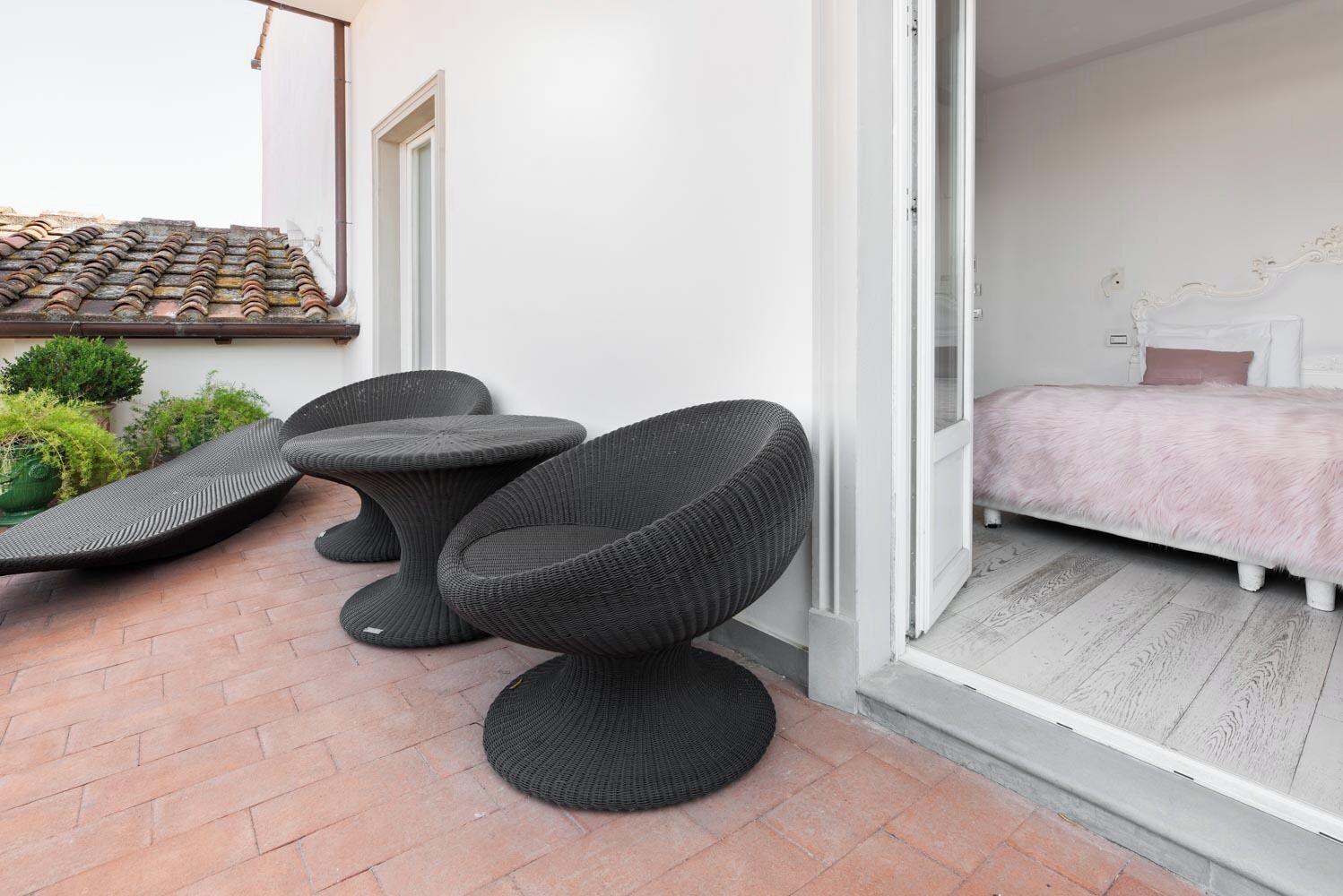 A modern outdoor seating area with wicker furniture, a tiled roof, and a view into a bedroom with a bed and white headboard.