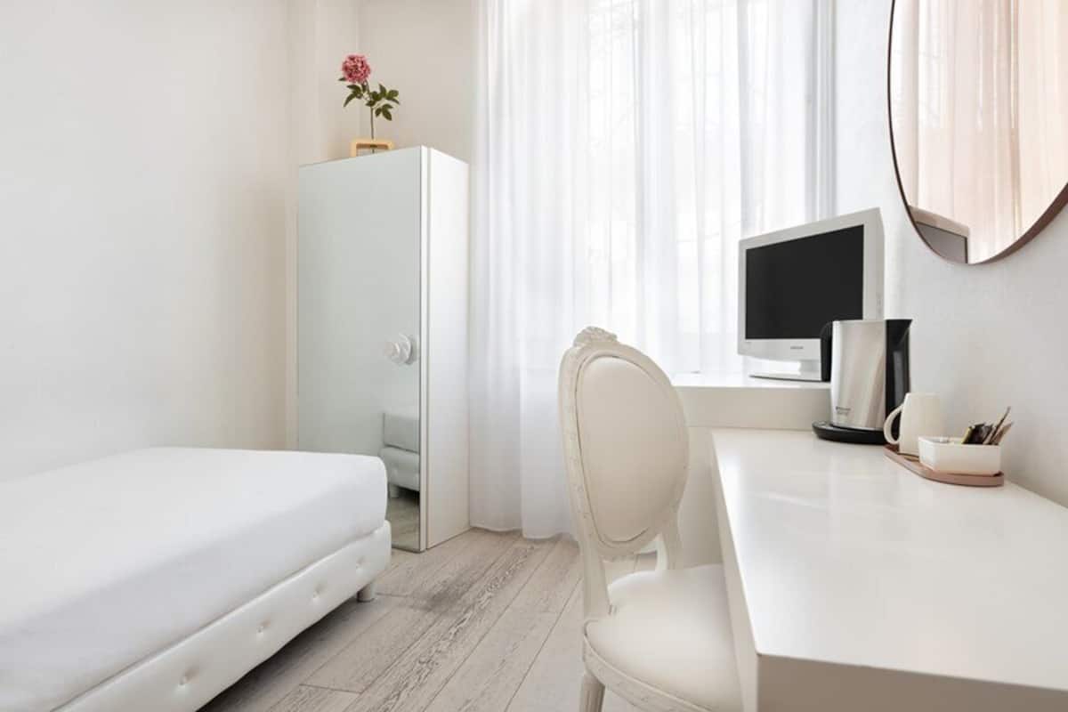 A minimalist hotel room with a bed, a desk, a chair, a computer, and a mirror.