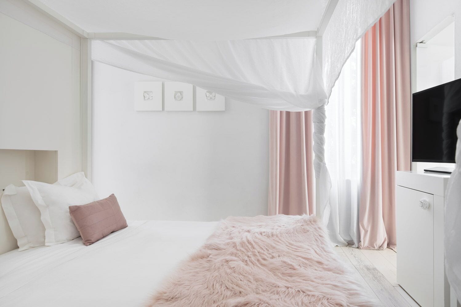 A modern bedroom with a large bed, a fluffy pink throw, white bedding, a TV on a cabinet, and sheer curtains.