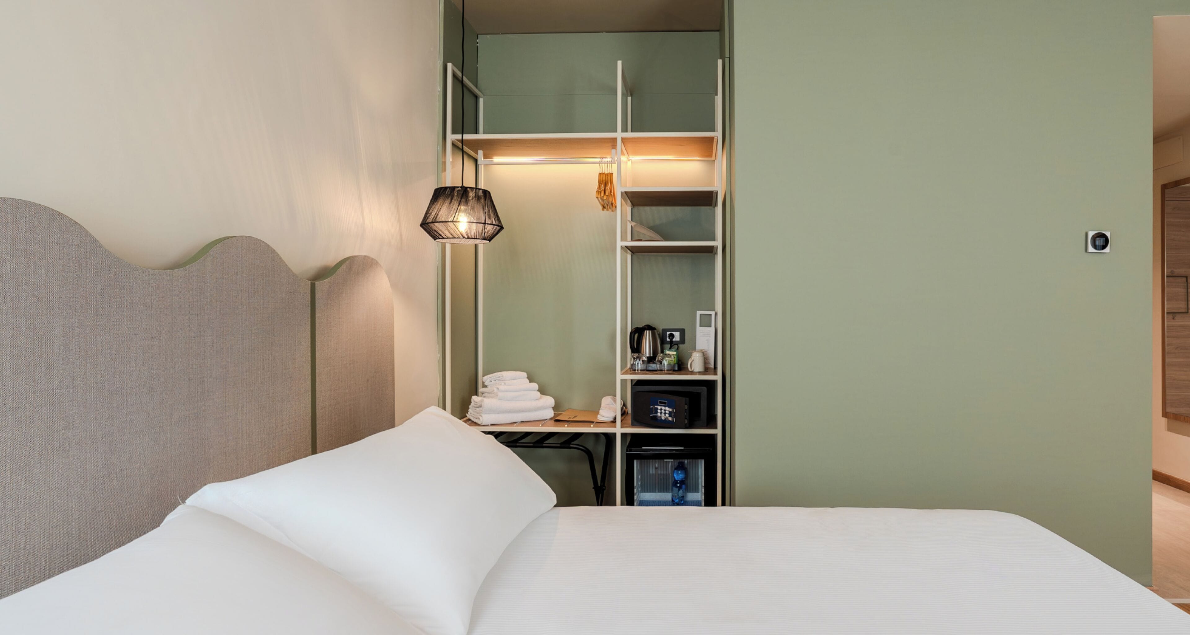A hotel room with a bed, a bedside lamp, a wardrobe, and a small table with a kettle.