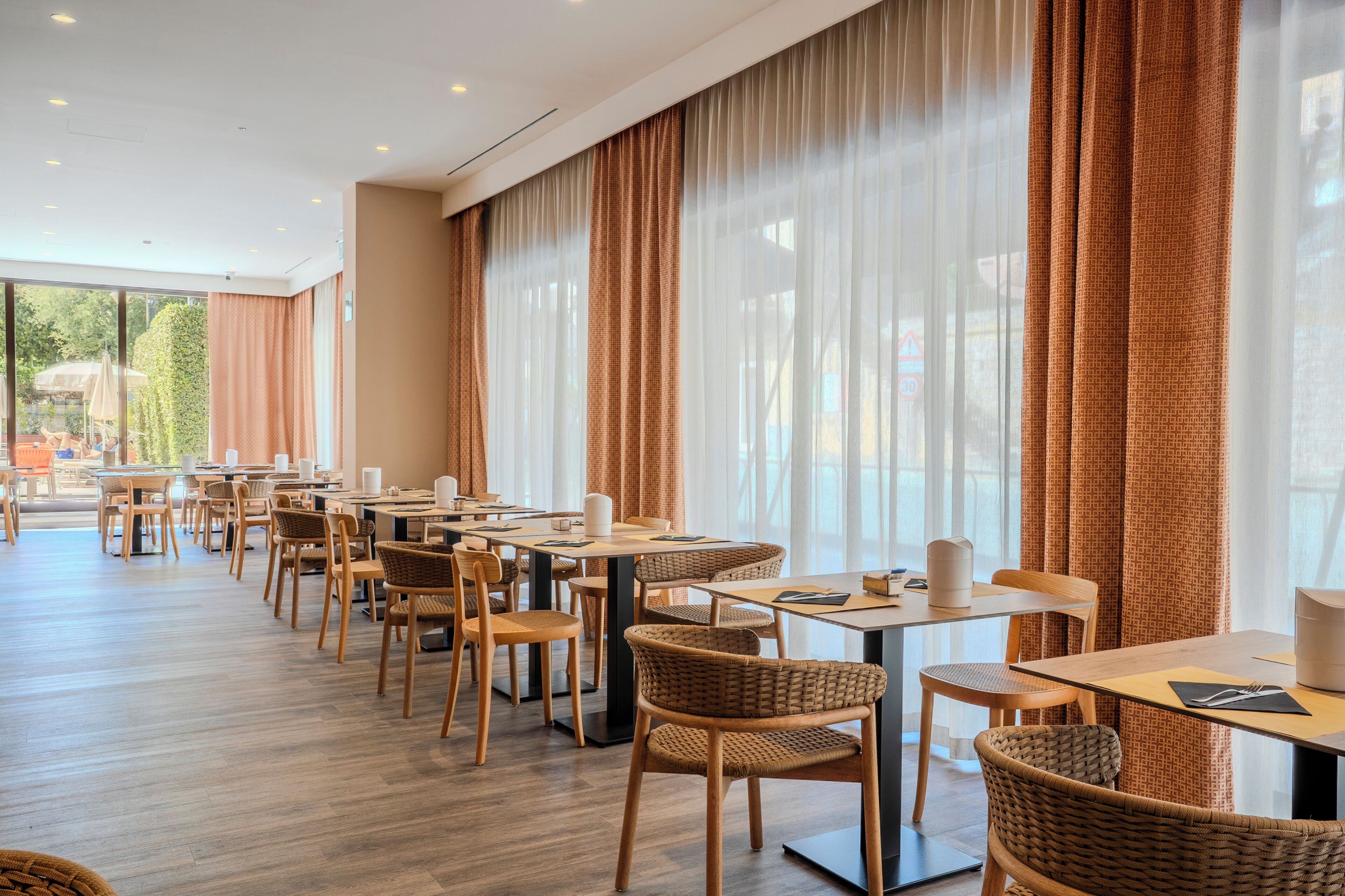 Um restaurante com várias mesas e cadeiras, janelas amplas com cortinas e vista para o exterior.