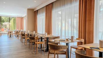 Um restaurante com várias mesas e cadeiras, janelas amplas com cortinas e vista para o exterior.