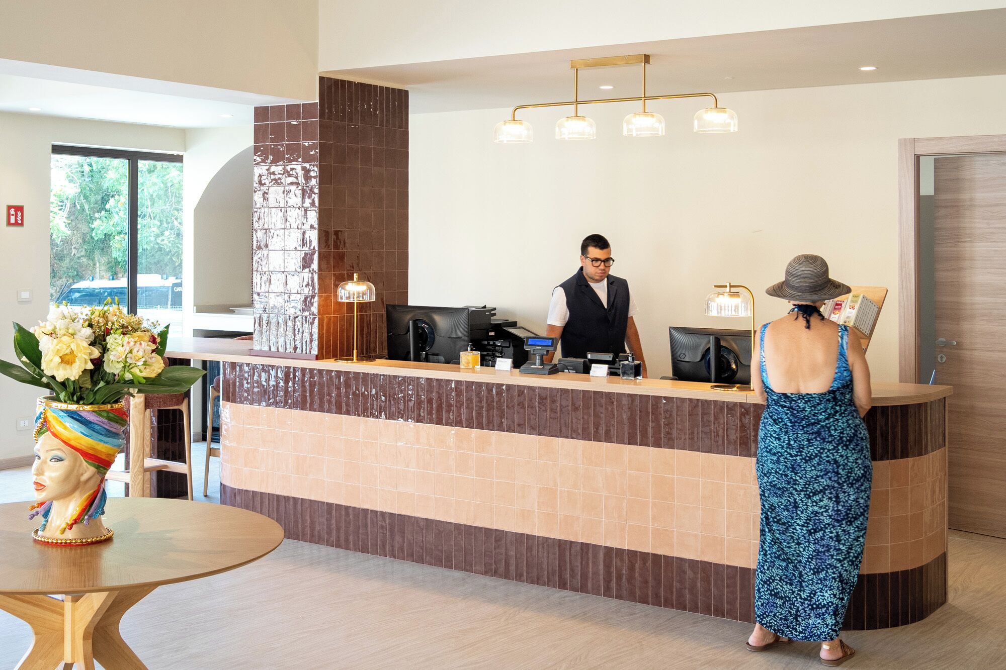A hotel reception desk with a man and a woman, a vase with flowers, and a table with a sculpture.