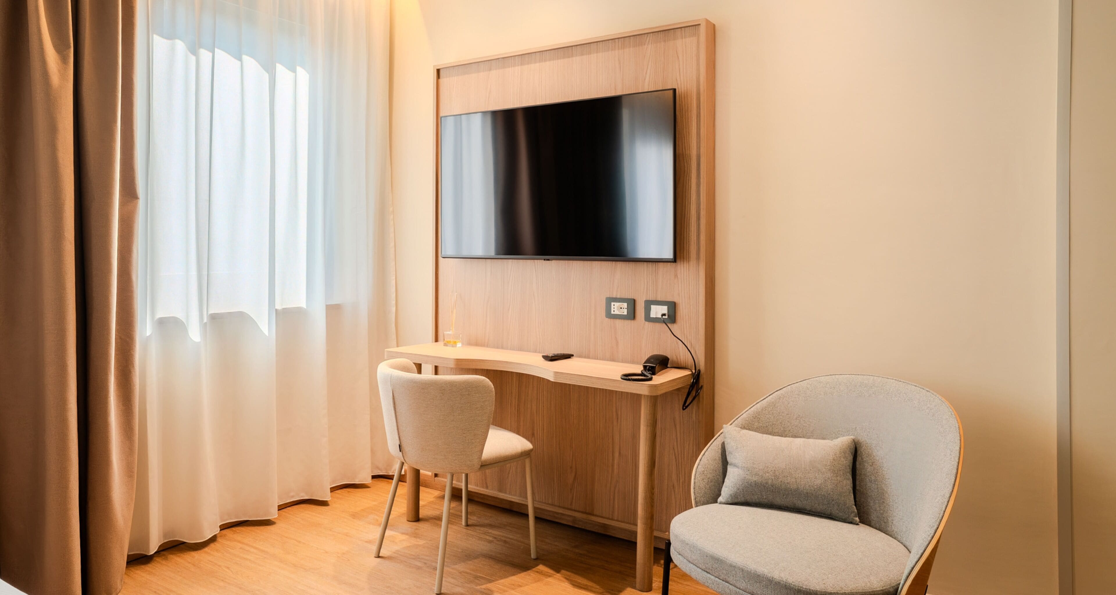 A modern hotel room with a flat-screen TV mounted on the wall, a wooden desk with a chair, and a cushioned armchair.