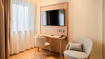 A modern hotel room with a flat-screen TV mounted on the wall, a wooden desk with a chair, and a cushioned armchair.