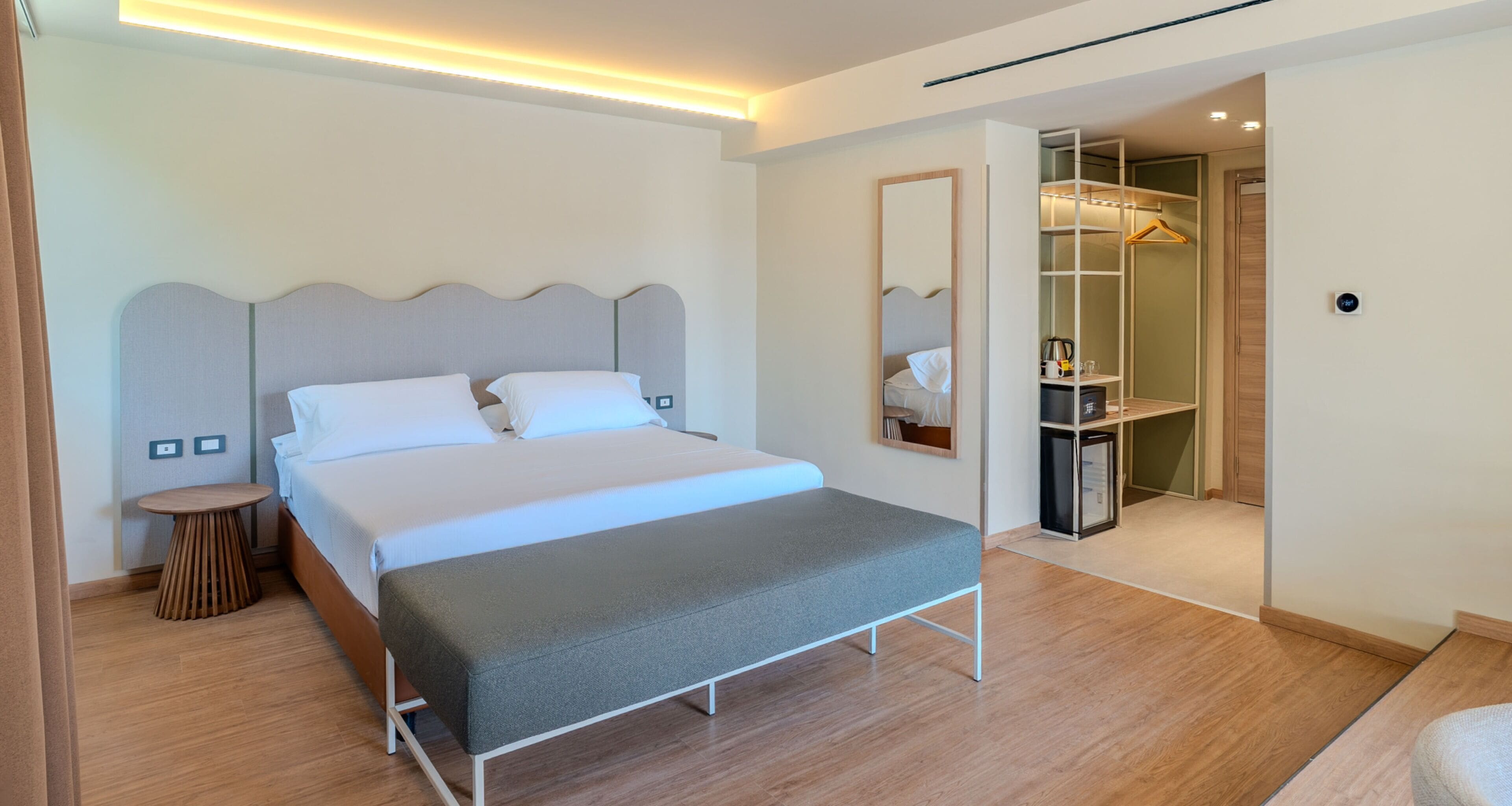 Una habitación de hotel moderna con una cama grande, mesitas de noche, un espejo y un vestidor.