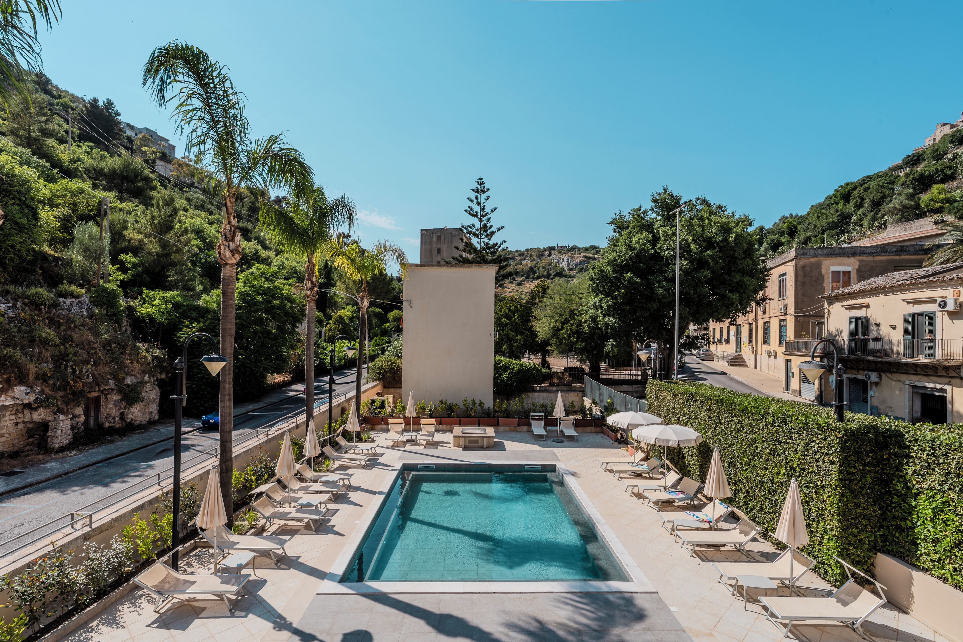 A swimming pool with lounge chairs and umbrellas, surrounded by greenery and buildings.