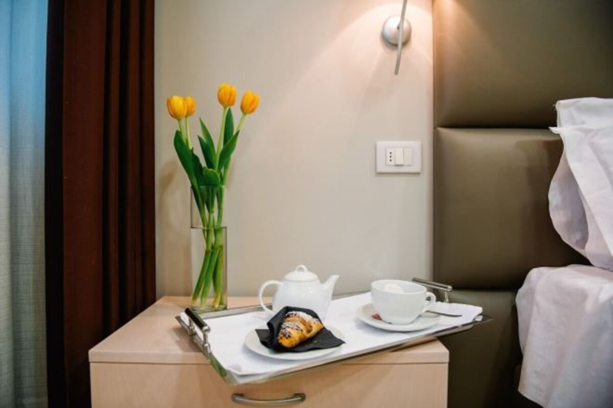 Una bandeja de desayuno ordenada con un croissant, una tetera y una taza, junto con un jarrón con tulipanes amarillos sobre una mesita de noche en una habitación de hotel.
