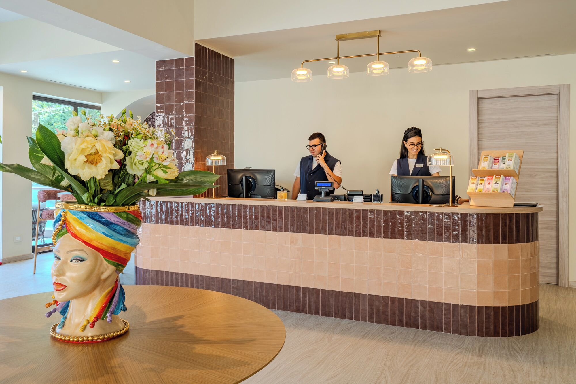 Et hotellets reception område med en buet disk, to receptionister, en vase med blomster og en kaffemaskine.