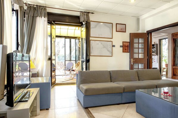 Ein modernes Wohnzimmer mit Sofa, Couchtisch und Fernseher. Vorhänge, gerahmte Bilder an der Wand und eine Tür zu einem anderen Raum sind zu sehen.