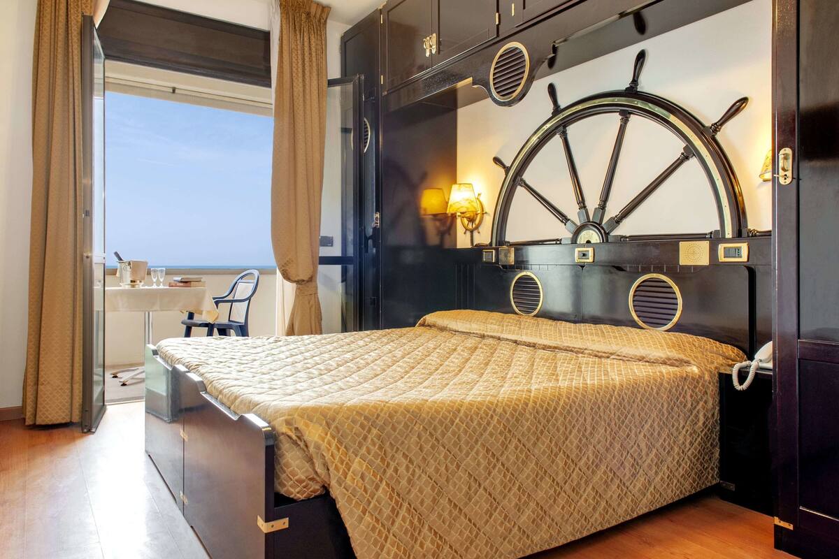 Un dormitorio con cama, escritorio y silla, un ventanal grande y un adorno con timón de barco.