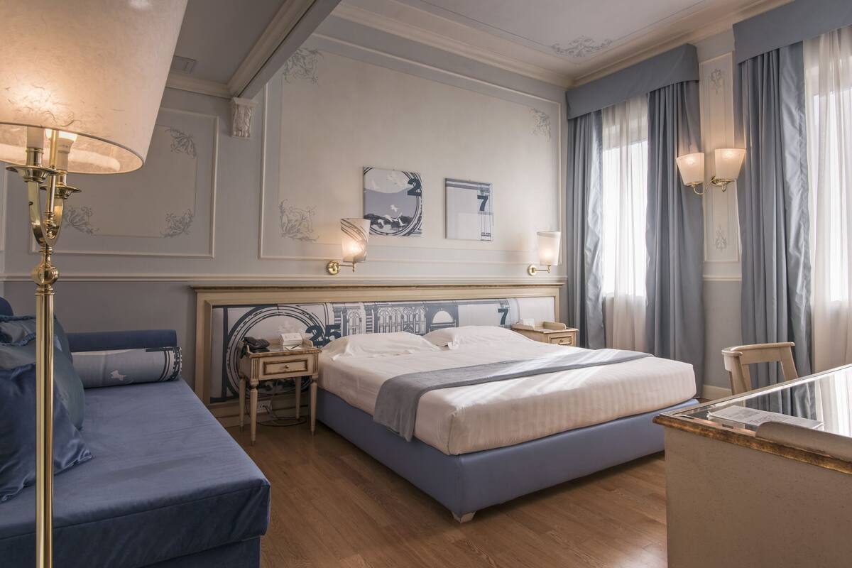 A hotel room with a large bed, a desk, a chair, and a nightstand with a lamp.