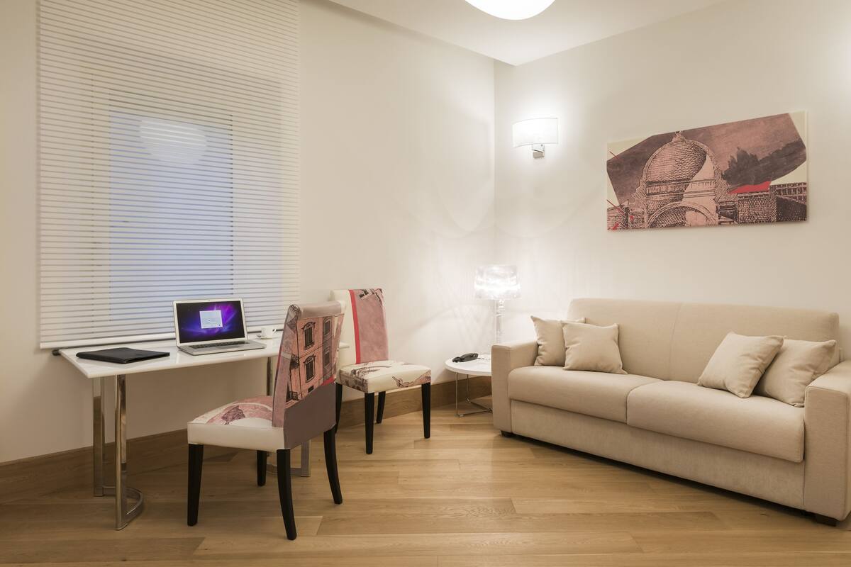 A modern living room with a white sofa, a desk with a laptop, a chair, a small table, and a wall-mounted lamp.