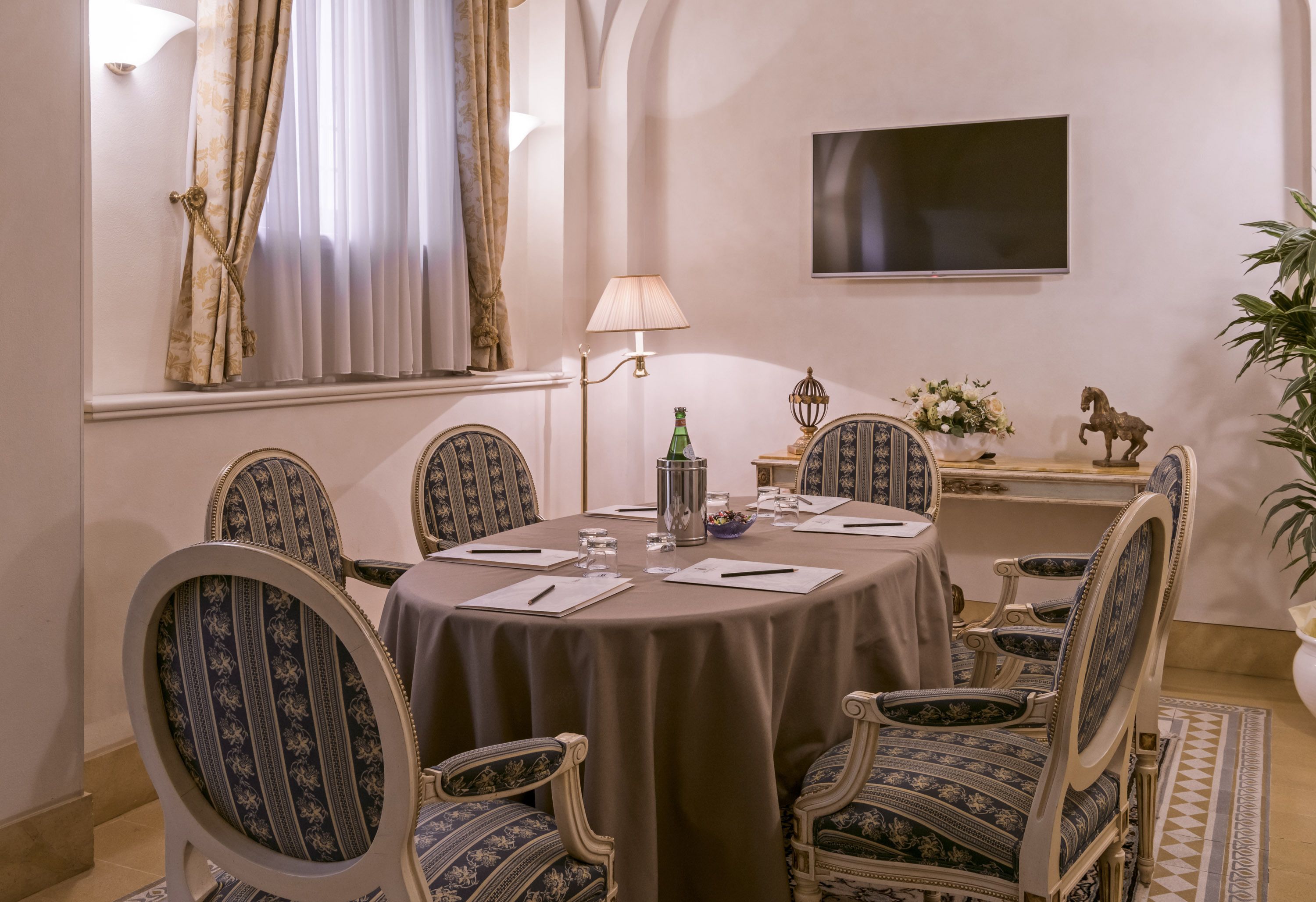 Une salle à manger élégante, avec une table ronde, six chaises rembourrées, un téléviseur à écran plat fixé au mur et une petite table d’appoint sur laquelle est posé un vase de fleurs.