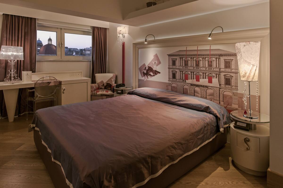 A hotel room with a large bed, a desk, a chair, a nightstand, and a view of buildings through the window.