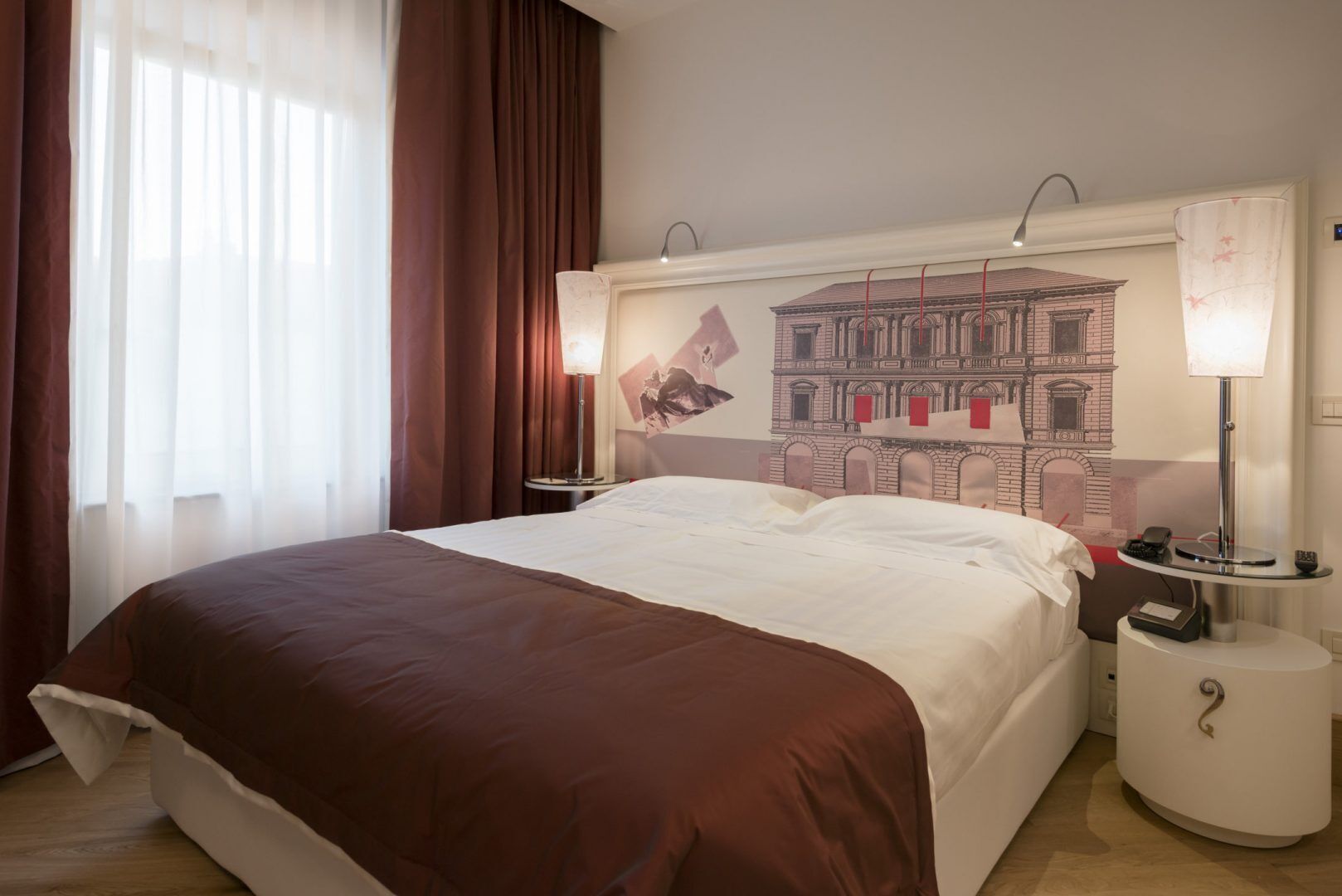 Quarto de hotel com uma cama grande, duas mesas de cabeceira, um criado-mudo, uma luminária na parede e uma janela com cortinas.