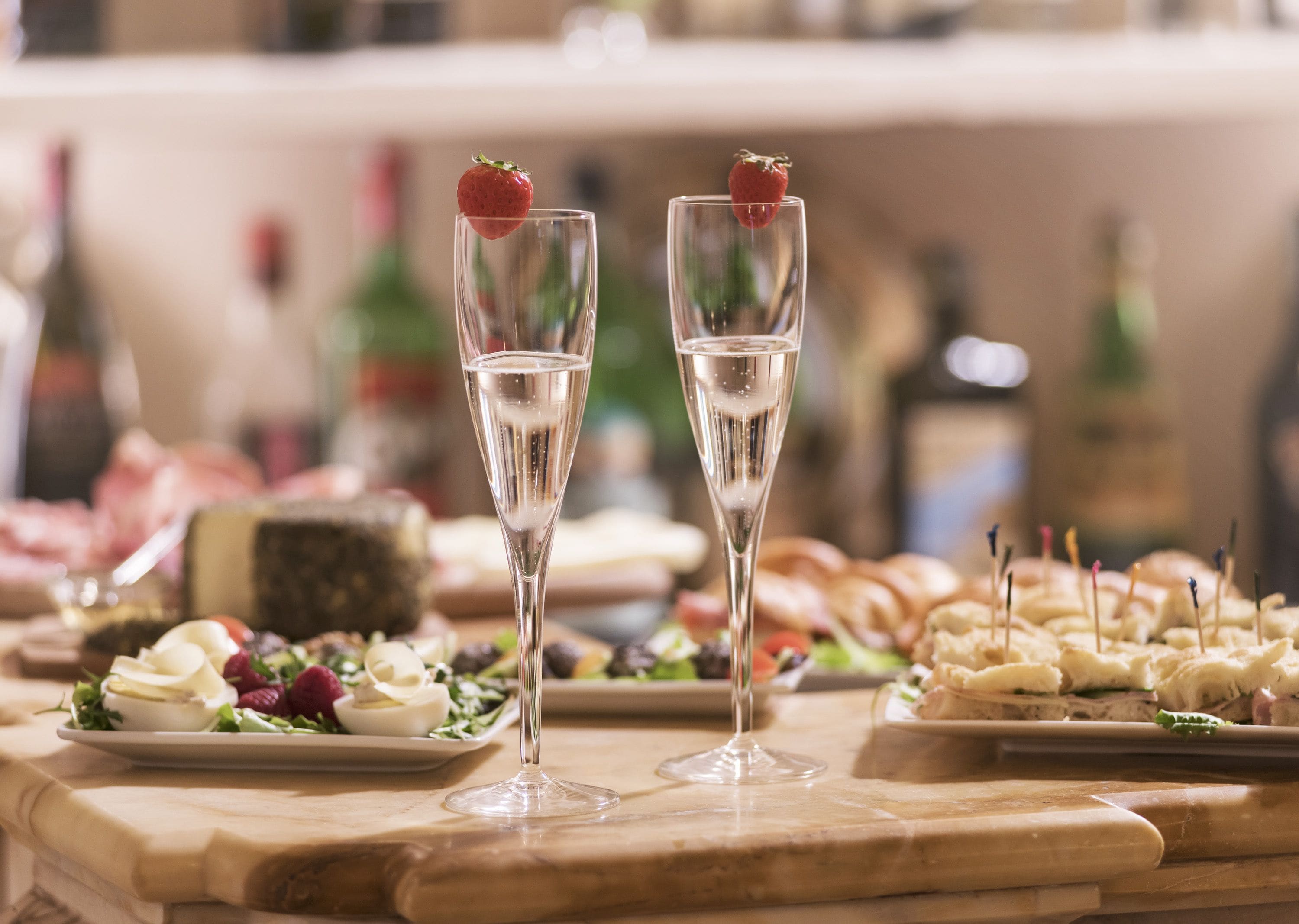 Two champagne flutes with strawberries on the rim, a variety of appetizers, and a cheese platter on a wooden table.