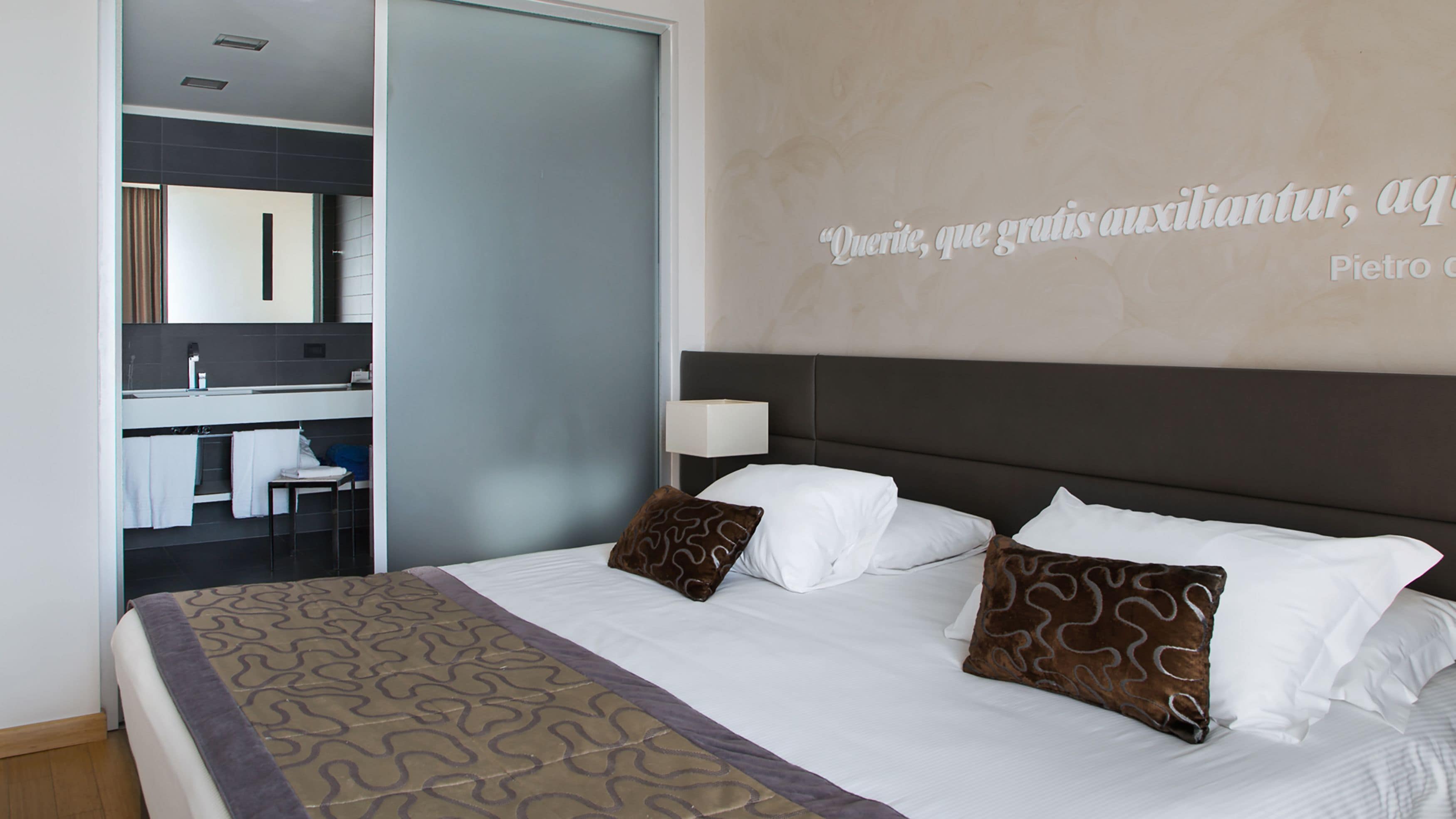 Een hotelkamer met een bed, nachtkastje, lamp, badkamer met wastafel en spiegel, en een handdoekhouder.