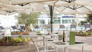 Een terras met witte tafels en stoelen, parasols, en zicht op een zwembad en gebouw op de achtergrond.
