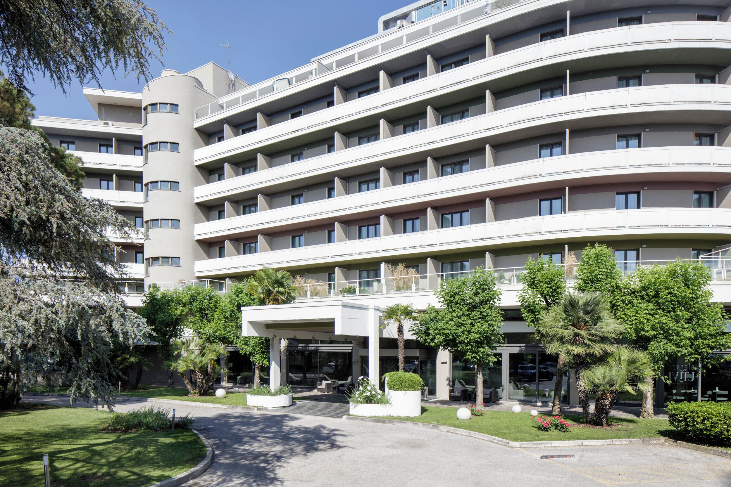 A multi-story hotel with a curved facade, balconies, and a landscaped entrance area with trees and shrubs.