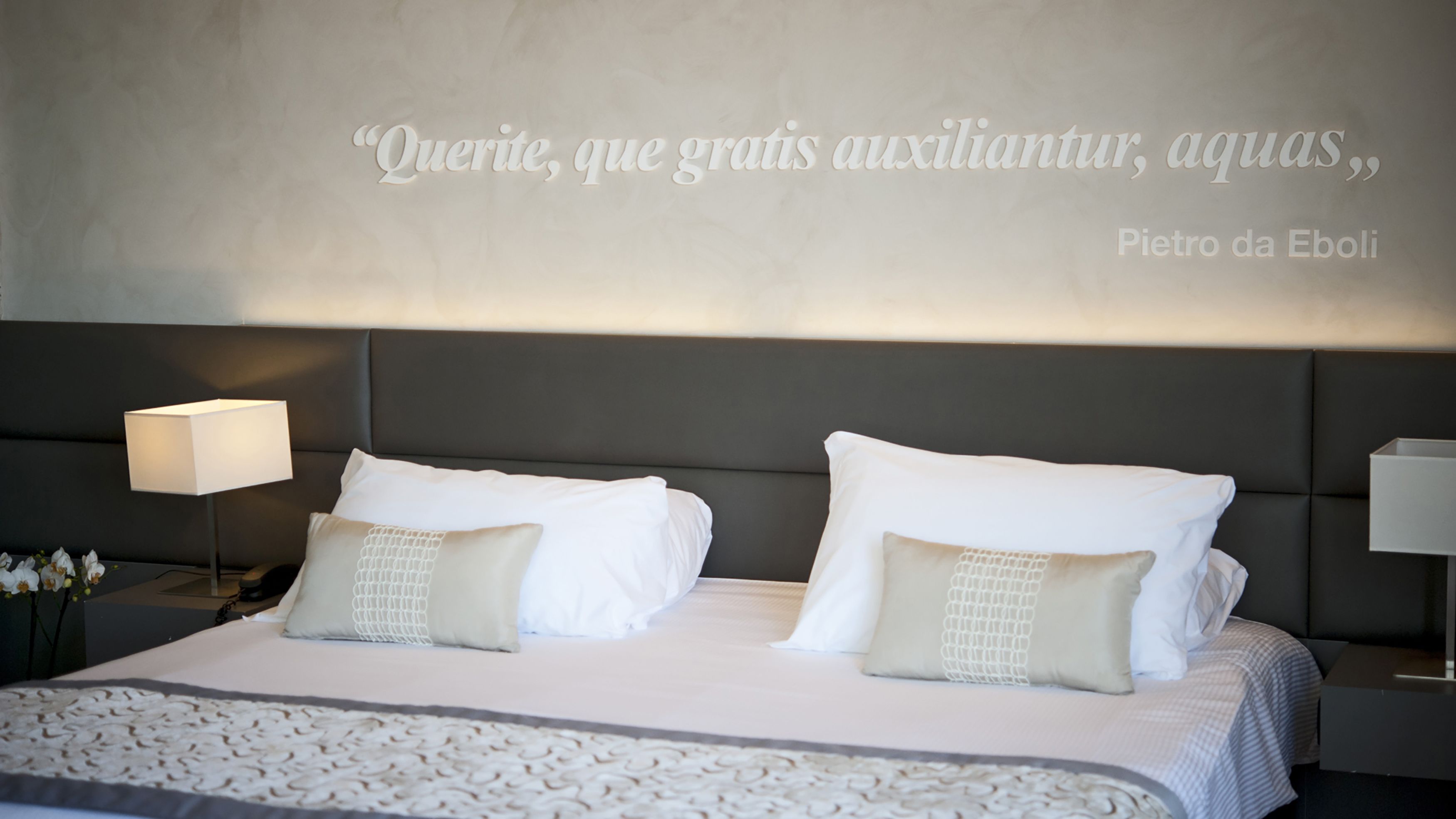 Ein ordentlich bezogenes Bett mit weißen Leinen und dekorativen Kissen, ein modernes Kopfbrett und eine Wand mit einem Zitat auf Portugiesisch.