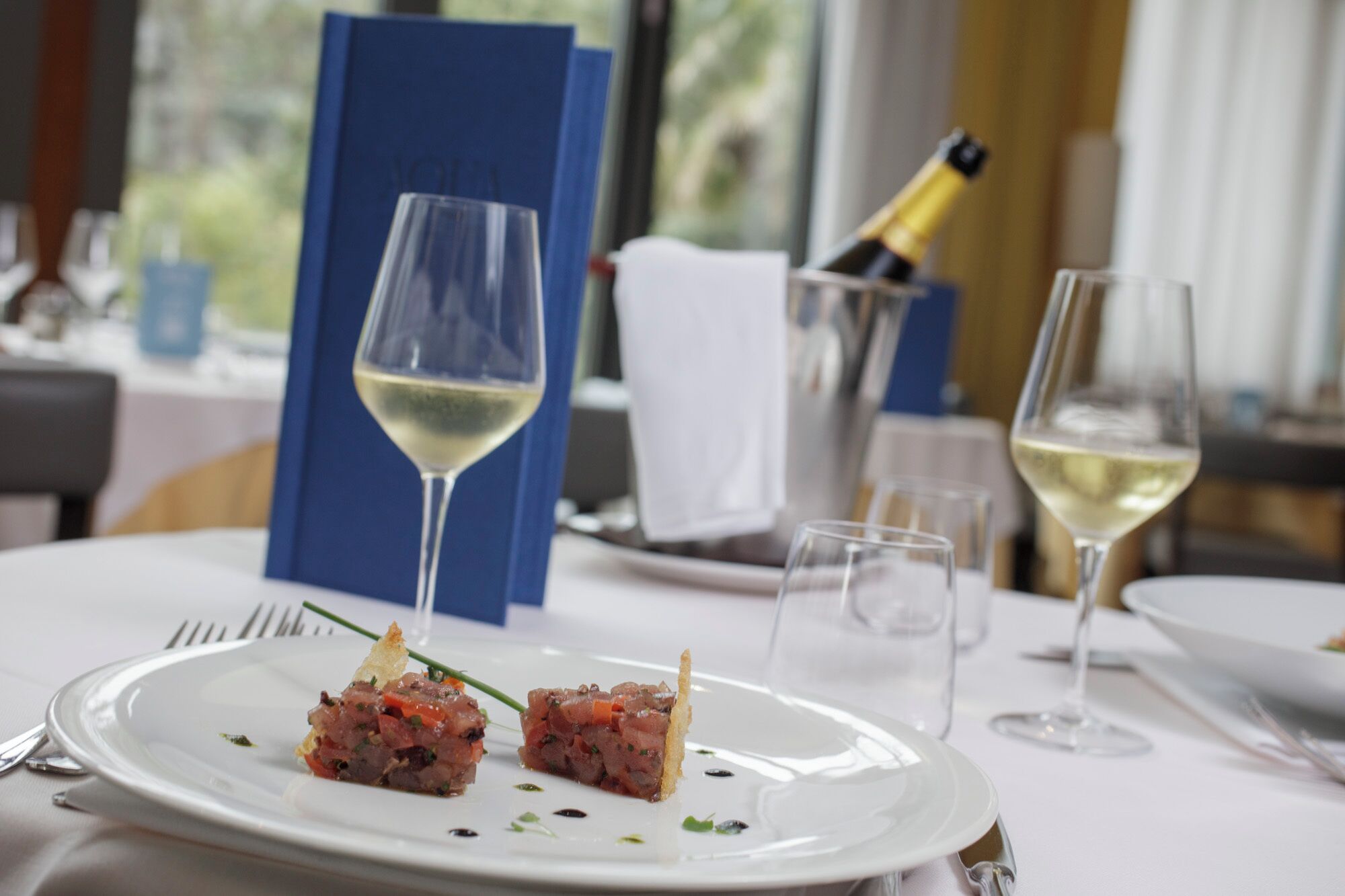 A dining table with a white plate of food, two wine glasses, and a bottle of champagne.