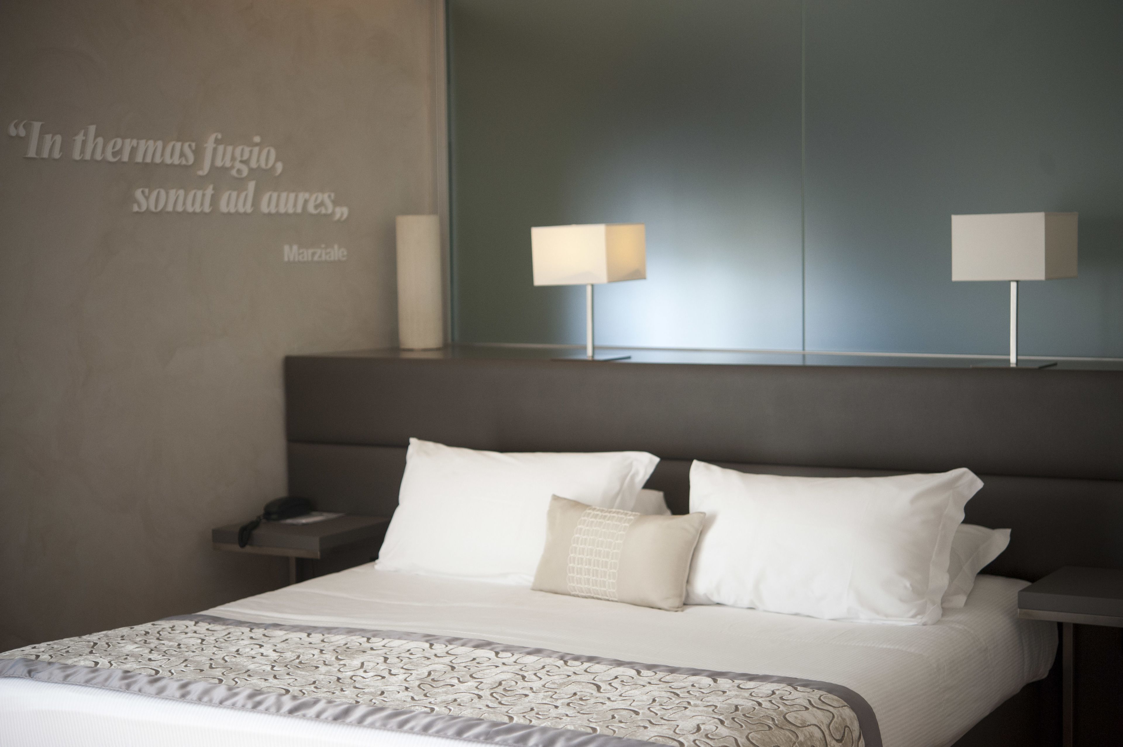 A modern hotel room with a large bed, two bedside lamps, a bedside table, and a wall quote.