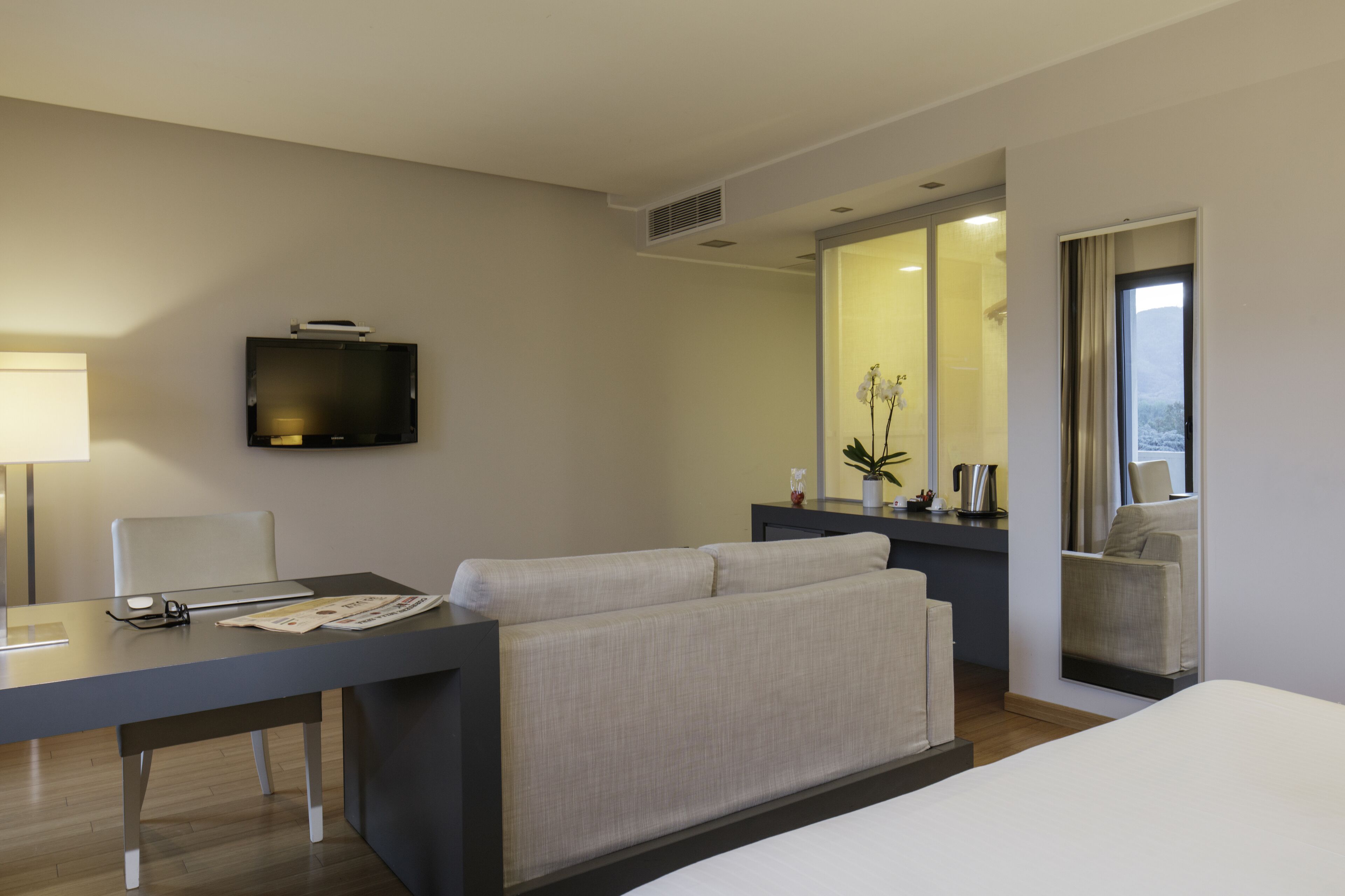 Ein modernes Hotelzimmer mit einem Bett, einem Sofa, einem Schreibtisch mit Computer, einem Fernseher und einem großen Spiegel, der den Raum reflektiert.