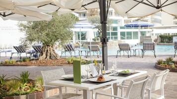 An outdoor dining area with white tables and chairs, a tree, and umbrellas.