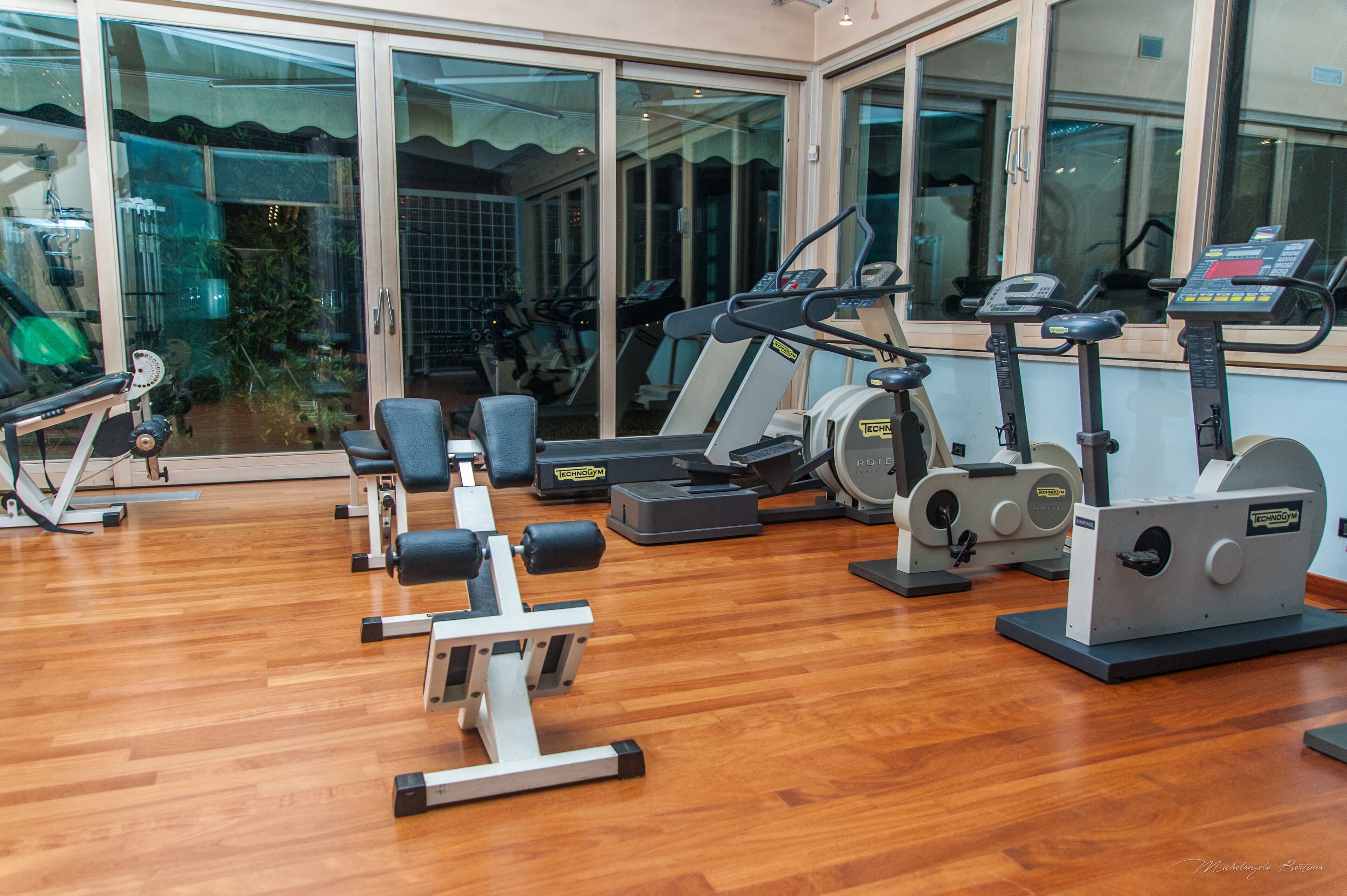 Un gimnasio con diversas máquinas de ejercicio, suelo de madera y grandes ventanales.