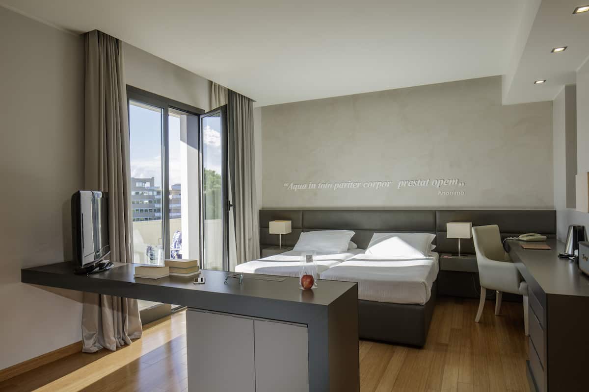 A modern hotel room with a large bed, a desk, and a view of the city through the window.